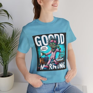 Könnte beinhalten: Ein hellblaues T-Shirt mit einem Grafikdruck eines Zombies im Schlafanzug, der eine Tasse Kaffee hält. Der Text "GOOD MORNING" ist in schwarzen Buchstaben über dem Zombie gedruckt und "GOOD MORNING." ist in einer Sprechblase über dem Kopf des Zombies gedruckt.