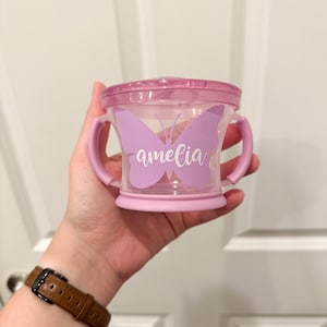 Pode incluir: Um copo de aprendizagem de pl&aacute;stico roxo com duas al&ccedil;as e um design de borboleta. A borboleta tem o nome "amelia" escrito nela.