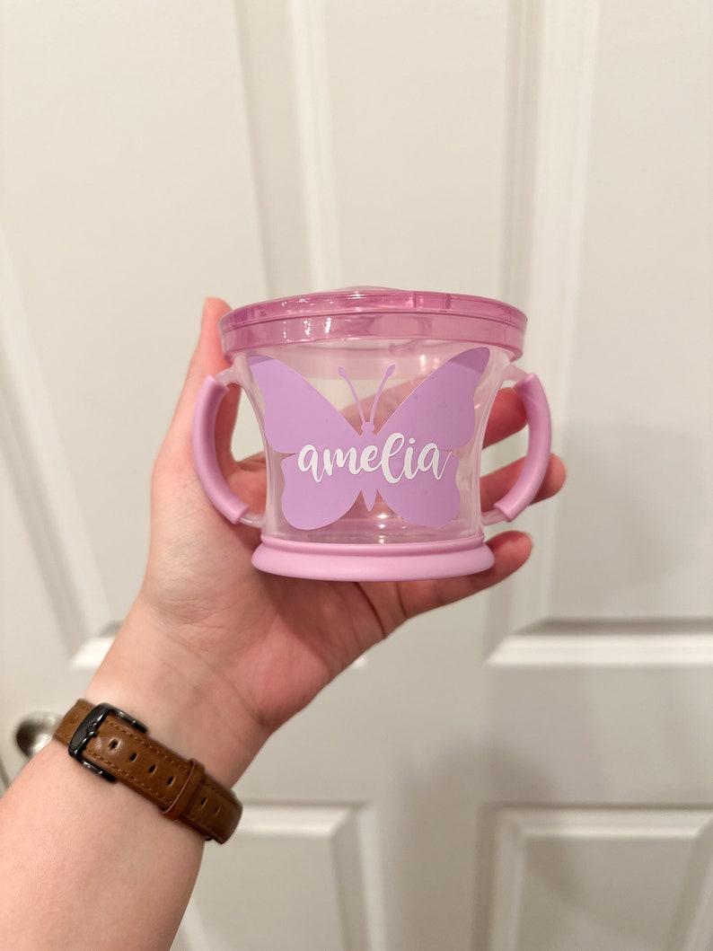 Pode incluir: Um copo de aprendizagem de pl&aacute;stico roxo com duas al&ccedil;as e um design de borboleta. A borboleta tem o nome "amelia" escrito nela.