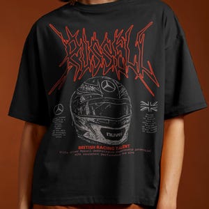 Puede incluir: Camiseta negra con un diseño gráfico rojo que presenta un casco de carreras y texto. La camiseta tiene mangas cortas y un ajuste holgado. La modelo lleva pantalones cortos marrones.