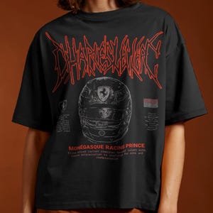 May include: A black short-sleeve t-shirt featuring a red graphic of a racing helmet and the text "CHARLES LECLERC" in a stylized font. Below the helmet, the text reads "MON&Eacute;GASQUE RACING PRINCE" in red, accompanied by smaller white text details.