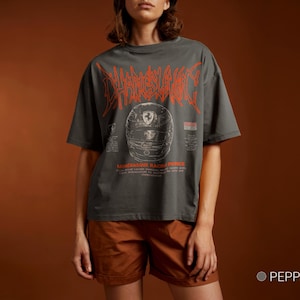 May include: A charcoal gray short-sleeve t-shirt featuring a large orange stylized graphic of a racing helmet with the text "CHARLES LECLERC" above it and "MONEGASQUE RACING PRINCE" below, along with smaller printed details on the front.
