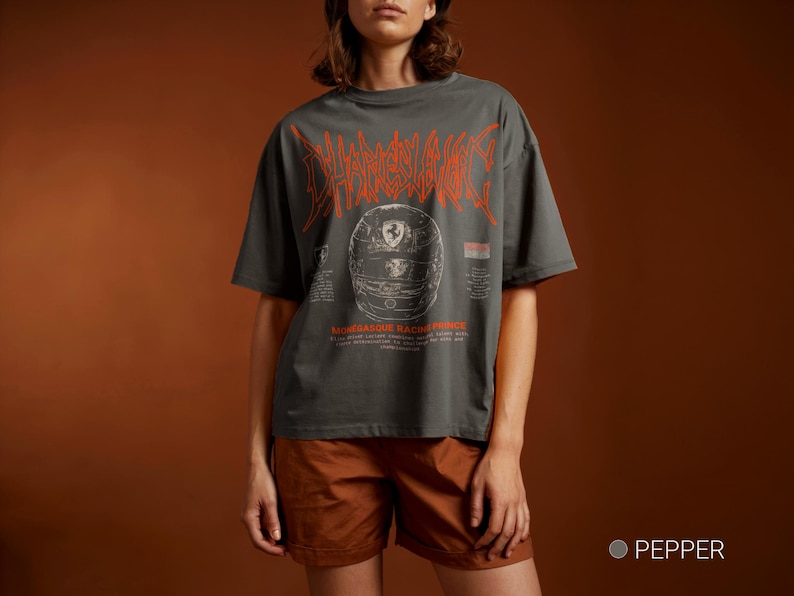 May include: A charcoal gray short-sleeve t-shirt featuring a large orange stylized graphic of a racing helmet with the text "CHARLES LECLERC" above it and "MONEGASQUE RACING PRINCE" below, along with smaller printed details on the front.