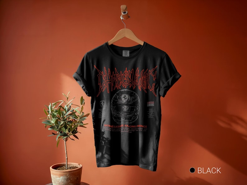 May include: A black short-sleeve t-shirt featuring a red graphic of a racing helmet and the text Charles Leclerc and Monegasque Racing Prince. The shirt has a crew neckline and rolled sleeves, displayed on a wooden hanger against a solid wall.