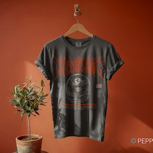 May include: A charcoal gray short-sleeve t-shirt featuring a graphic of a racing helmet in the center with orange stylized text above it. The design includes smaller text and a small logo below the helmet graphic. The shirt is displayed on a hanger.
