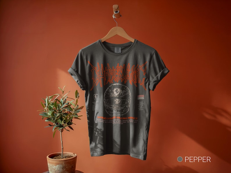 May include: A charcoal gray short-sleeve t-shirt featuring a graphic of a racing helmet in the center with orange stylized text above it. The design includes smaller text and a small logo below the helmet graphic. The shirt is displayed on a hanger.