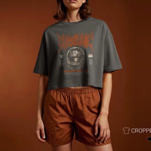 May include: A charcoal gray cropped boxy t-shirt featuring an orange graphic of a racing helmet with stylized text above it. The design includes small logos and text details on either side of the helmet. The shirt is paired with brown shorts.