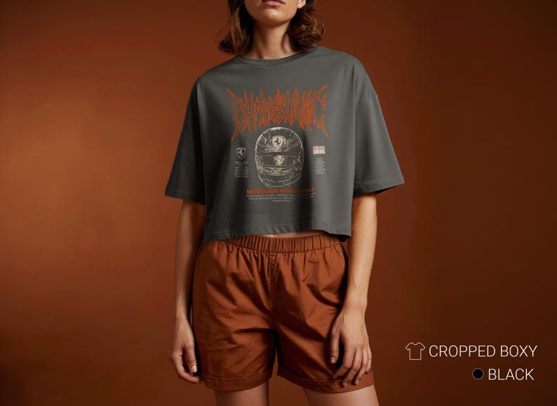 May include: A charcoal gray cropped boxy t-shirt featuring an orange graphic of a racing helmet with stylized text above it. The design includes small logos and text details on either side of the helmet. The shirt is paired with brown shorts.