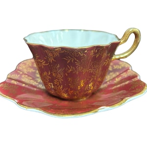 May include: A red and gold teacup and saucer set with a floral pattern. The cup and saucer have a scalloped edge.