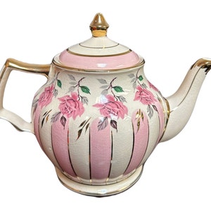 Könnte beinhalten: Eine weiße Teekanne mit rosa und goldenen Streifen und floralen Akzenten. Die Teekanne hat einen Deckel mit einem goldenen Knopf und einen Ausguss an der Seite.