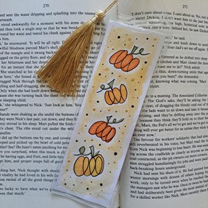 Könnte beinhalten: Handgefertigter Lesezeichen mit goldener Quaste, mit einem Aquarelldesign von drei orangefarbenen Kürbissen mit grünen Stielen. Das Lesezeichen befindet sich in einem offenen Buch.