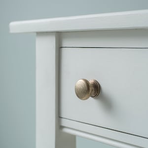 May include: White wooden drawer with a single brass knob. The drawer is part of a white wooden bedside table.