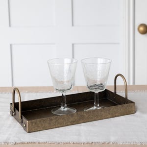 May include: A rectangular, antique brass tray with two clear stemmed glasses. The tray has curved handles and is set on a neutral-colored table runner. The glasses have a delicate floral pattern.