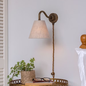 May include: A wall-mounted bedside lamp with a beige fabric shade and a curved, antique brass arm. The lamp is connected to a wall outlet with a matching cord. A small table with a plant and glasses is below the lamp.