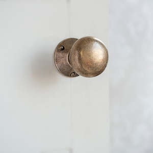May include: A round, brass door knob with a slightly aged finish. The knob is attached to a round, brass plate with two visible screws.