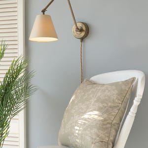 May include: A brass wall lamp with a beige shade is positioned above a white chair. A cushion with a floral design sits on the chair. The wall is painted a pale blue. The lamp's arm is adjustable.