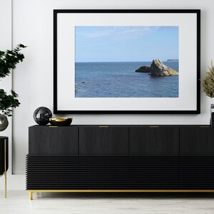 Op de afbeelding: Een zwart ingelijst beeld met een foto van een rotsachtig eiland in de oceaan. De afbeelding hangt aan een witte muur boven een zwarte commode met gouden accenten.
