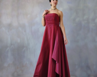May include: A woman wearing a strapless, burgundy-colored gown with a flowing, asymmetrical skirt. The gown has a simple design and is made of a smooth, silky fabric.