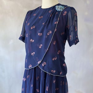 Ocean blue vintage chiffon dress from the 70s with a dust pink glitter leaf print and sequin detail at the bust. Features include a silver-trimmed wrap top, sheer chiffon sleeves with pink pearls, and a softly pleated A-line midi skirt. Fully lined in burgundy satin for comfort. A delicate and stylish piece ideal for wedding guests, spring events, and summer dinners. In perfect condition, recommended for small to medium sizes.