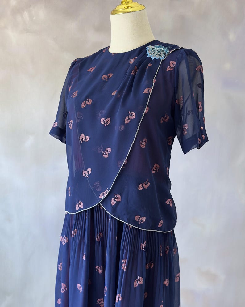 Ocean blue vintage chiffon dress from the 70s with a dust pink glitter leaf print and sequin detail at the bust. Features include a silver-trimmed wrap top, sheer chiffon sleeves with pink pearls, and a softly pleated A-line midi skirt. Fully lined in burgundy satin for comfort. A delicate and stylish piece ideal for wedding guests, spring events, and summer dinners. In perfect condition, recommended for small to medium sizes.