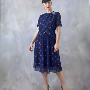 Ocean blue vintage chiffon dress from the 1970s with a dust pink glitter leaf print and light blue sequin appliqué on the chest. Features a silver-trimmed wrap bodice, round neckline, and sheer short sleeves finished with pink-toned pearls. The A-line midi skirt is softly pleated at the waistband and flows below. Fully lined in burgundy satin for contrast. Perfect for weddings, garden parties, and summer dinners. Romantic and elegant, in flawless vintage condition. Fits small to medium.