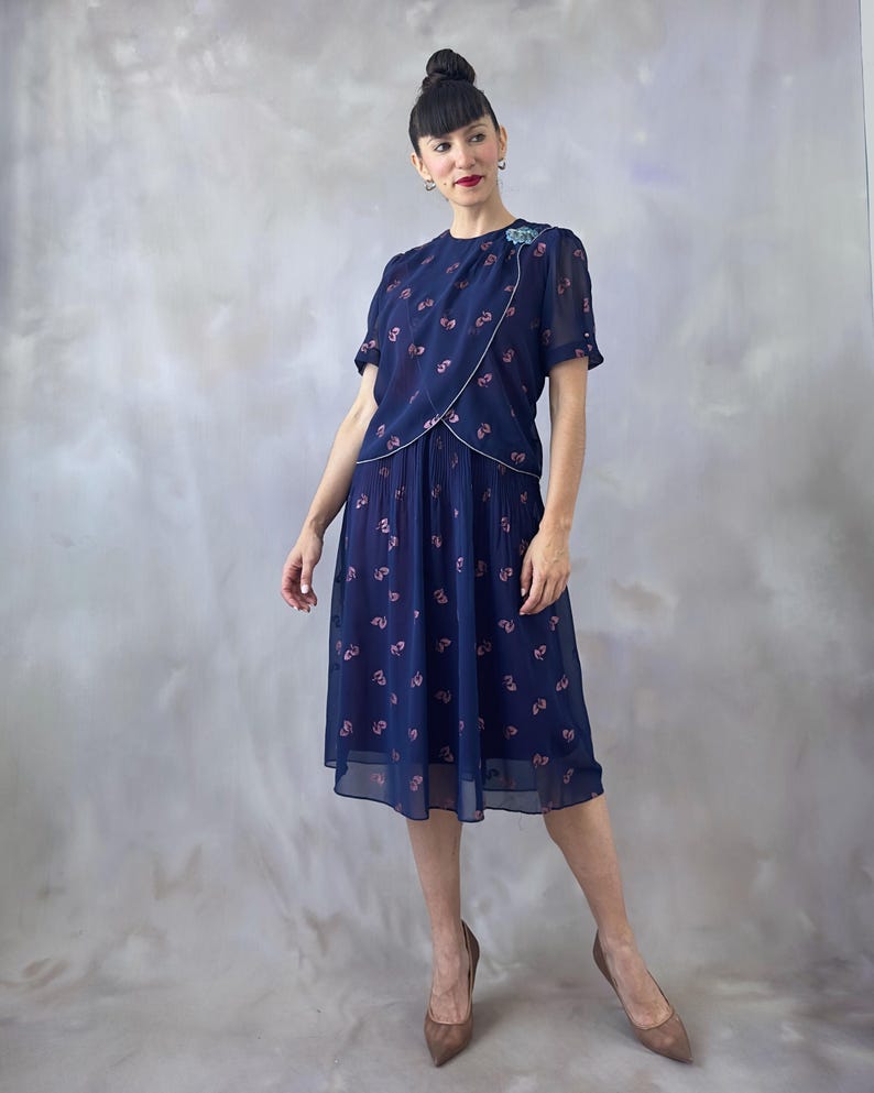 Ocean blue vintage chiffon dress from the 1970s with a dust pink glitter leaf print and light blue sequin appliqué on the chest. Features a silver-trimmed wrap bodice, round neckline, and sheer short sleeves finished with pink-toned pearls. The A-line midi skirt is softly pleated at the waistband and flows below. Fully lined in burgundy satin for contrast. Perfect for weddings, garden parties, and summer dinners. Romantic and elegant, in flawless vintage condition. Fits small to medium.