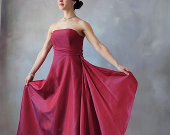 May include: A woman wearing a strapless, crimson red, floor-length gown with a fitted bodice and a flowing skirt.