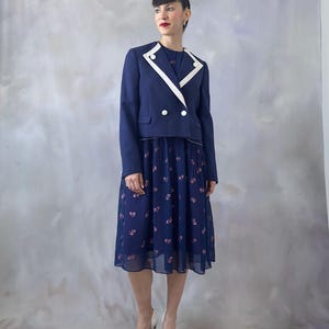 Chiffon 70s cocktail dress in deep ocean blue with pink glitter leaves and a sequin appliqué at the chest. Features a relaxed wrap-style bodice trimmed in silver, short sheer sleeves with pearl details, and an A-line skirt pleated at the top. Lined in contrasting burgundy satin for elegance and comfort. A timeless and lightweight vintage piece perfect for wedding guests, dinners, and special occasions. Best for size S/M.