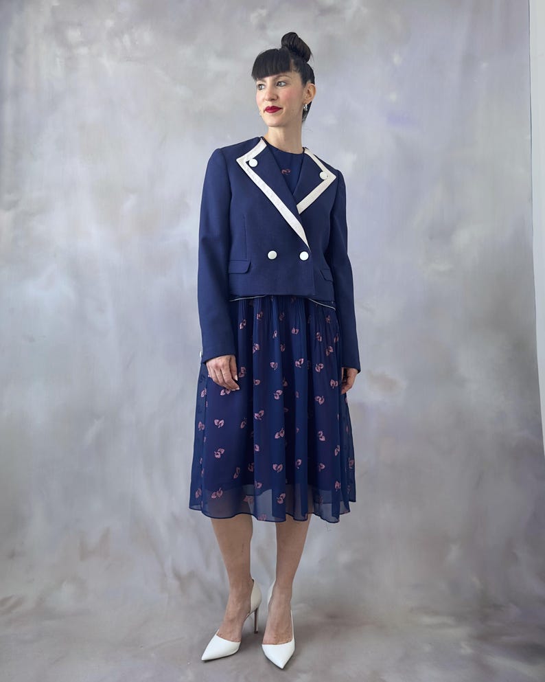 Chiffon 70s cocktail dress in deep ocean blue with pink glitter leaves and a sequin appliqué at the chest. Features a relaxed wrap-style bodice trimmed in silver, short sheer sleeves with pearl details, and an A-line skirt pleated at the top. Lined in contrasting burgundy satin for elegance and comfort. A timeless and lightweight vintage piece perfect for wedding guests, dinners, and special occasions. Best for size S/M.