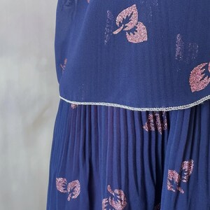 Ocean blue chiffon dress from the 1970s featuring a pink glitter leaf print and sequin detail on the chest. Silver-trimmed wrap-style bodice, round neckline, and sheer sleeves with pink-toned pearls. The A-line midi skirt is softly pleated at the top. Lined in burgundy satin for contrast and comfort. A graceful vintage option for wedding guests, spring events, and elegant dinners. Size S/M.