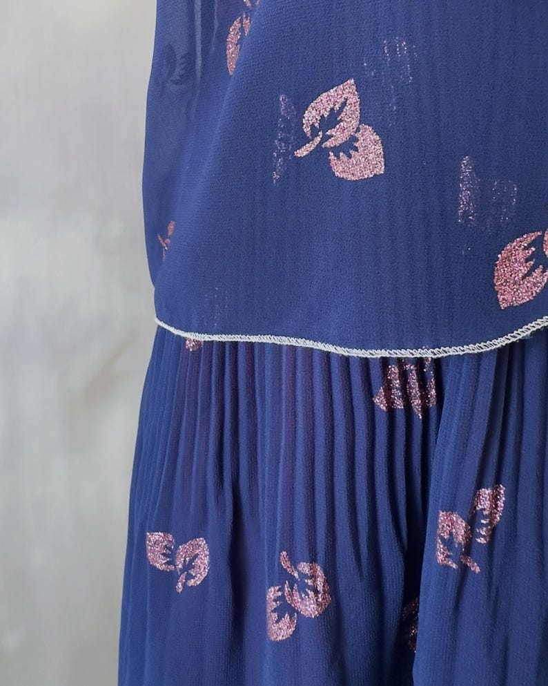 Ocean blue chiffon dress from the 1970s featuring a pink glitter leaf print and sequin detail on the chest. Silver-trimmed wrap-style bodice, round neckline, and sheer sleeves with pink-toned pearls. The A-line midi skirt is softly pleated at the top. Lined in burgundy satin for contrast and comfort. A graceful vintage option for wedding guests, spring events, and elegant dinners. Size S/M.