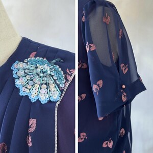 Romantic chiffon vintage dress from the 1970s in dark ocean blue with pink glitter leaf print and a sequin appliqué on the chest. Silver-trimmed wrap bodice, round neckline, and short sleeves with sheer chiffon and pink pearl detail. The A-line skirt falls to midi length and is softly pleated at the waist. Burgundy satin lining adds depth. Ideal for weddings, garden parties, and cocktail events. Best for size S/M.