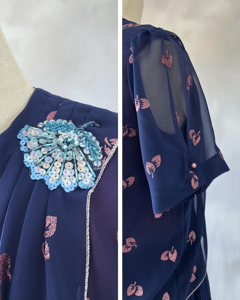 Romantic chiffon vintage dress from the 1970s in dark ocean blue with pink glitter leaf print and a sequin appliqué on the chest. Silver-trimmed wrap bodice, round neckline, and short sleeves with sheer chiffon and pink pearl detail. The A-line skirt falls to midi length and is softly pleated at the waist. Burgundy satin lining adds depth. Ideal for weddings, garden parties, and cocktail events. Best for size S/M.