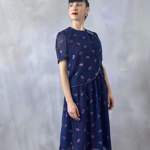 70s vintage chiffon dress in ocean blue with a pink glitter leaf pattern and sequin detail on the bodice. The wrap top is silver-trimmed with short transparent sleeves finished with pink pearls. The A-line skirt is pleated only at the waistband and flows to a midi length. Burgundy satin lining offers a soft contrast. Ideal for wedding guests, spring garden events, or formal summer occasions. In perfect condition and recommended for sizes S/M.