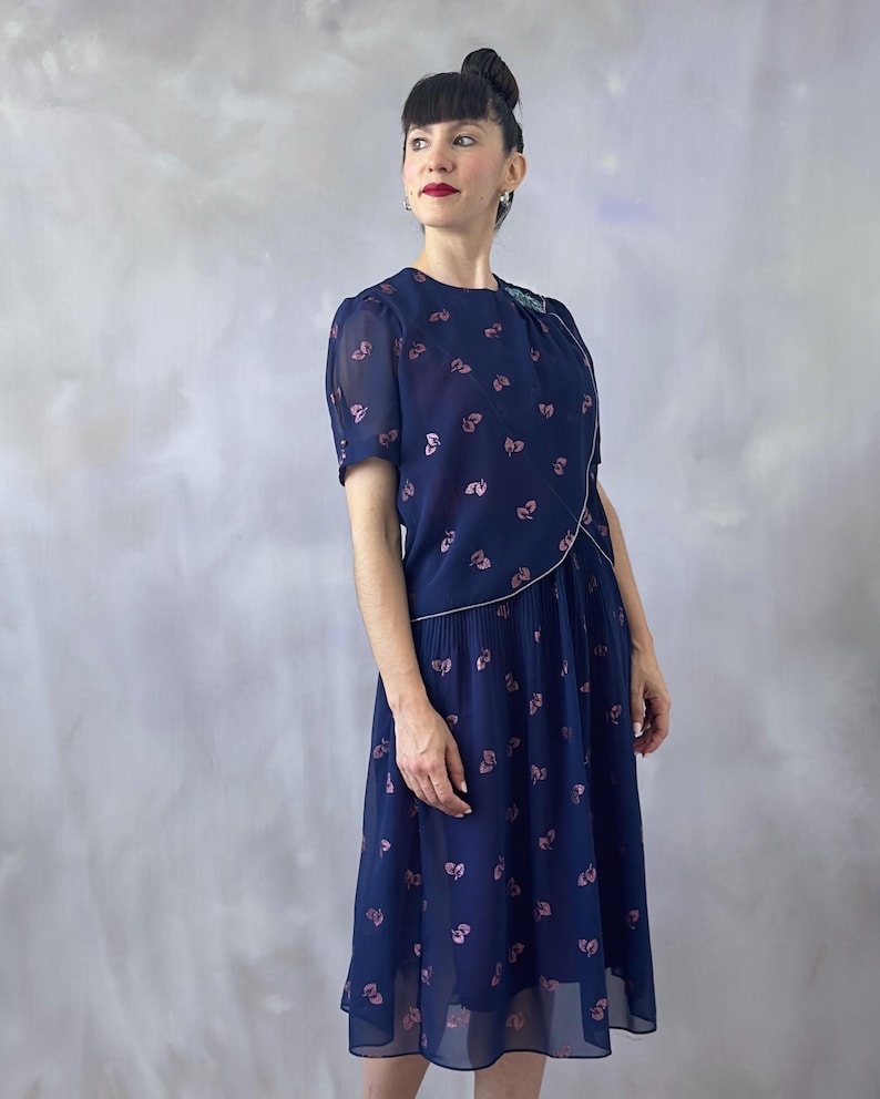 70s vintage chiffon dress in ocean blue with a pink glitter leaf pattern and sequin detail on the bodice. The wrap top is silver-trimmed with short transparent sleeves finished with pink pearls. The A-line skirt is pleated only at the waistband and flows to a midi length. Burgundy satin lining offers a soft contrast. Ideal for wedding guests, spring garden events, or formal summer occasions. In perfect condition and recommended for sizes S/M.