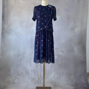 Elegant 1970s chiffon dress in ocean blue with glittery pink leaves printed throughout and a sequin chest detail. The wrap bodice has a silver trim, round neckline, and sheer sleeves finished with pearl accents. The A-line midi skirt is softly pleated at the top. Fully lined in burgundy satin. A flowy and romantic piece ideal for spring or summer weddings and garden parties. In excellent condition, fits size S/M.