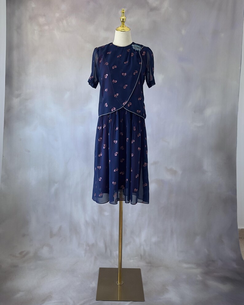 Elegant 1970s chiffon dress in ocean blue with glittery pink leaves printed throughout and a sequin chest detail. The wrap bodice has a silver trim, round neckline, and sheer sleeves finished with pearl accents. The A-line midi skirt is softly pleated at the top. Fully lined in burgundy satin. A flowy and romantic piece ideal for spring or summer weddings and garden parties. In excellent condition, fits size S/M.