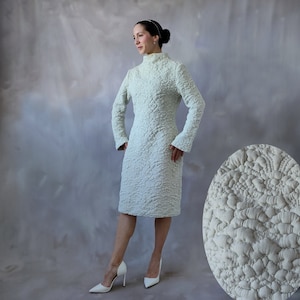 May include: A white dress with a textured, quilted pattern. The dress has a high neck and long sleeves. The model is wearing white heels.