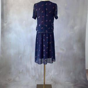 Chiffon 70s vintage dress in ocean blue with a pink glitter leaf print and sequin accent at the chest. Wrap bodice with silver trim, round neckline, and transparent sleeves finished with pink-toned pearls. A-line midi skirt pleated only at the waistband and fully lined with burgundy satin. Lightweight and elegant, perfect for spring weddings and evening dinners. Excellent condition, size S/M.
