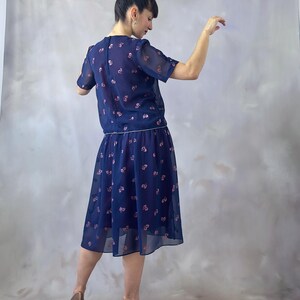 Puede incluir: Vestido de dos piezas azul marino con estampado floral. La parte superior es una blusa de manga corta y la falda llega a la rodilla. El vestido tiene un aspecto delicado y transparente. La modelo lleva tacones altos marrones.