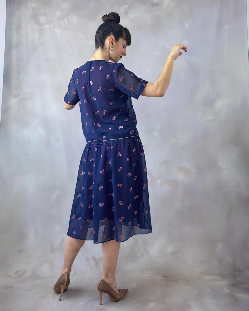 Puede incluir: Vestido de dos piezas azul marino con estampado floral. La parte superior es una blusa de manga corta y la falda llega a la rodilla. El vestido tiene un aspecto delicado y transparente. La modelo lleva tacones altos marrones.