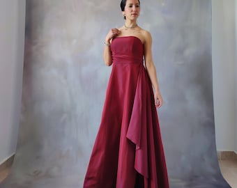 May include: A woman wearing a strapless, burgundy, floor-length gown with a draped skirt. The gown has a simple, elegant design.