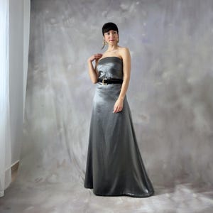 May include: A strapless, floor-length silver-gray gown with a fitted bodice and a flowing skirt. The dress is cinched at the waist with a black belt featuring a gold buckle. The fabric has a subtle sheen.