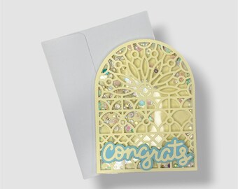 May include: A pale yellow arched shaker card featuring a laser-cut geometric lattice pattern filled with iridescent sequins and stars. The word "congrats" is written in a light blue script font across the bottom, accompanied by a plain white envelope.