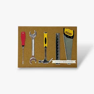 May include: A rectangular brown greeting card featuring a collection of miniature tool embellishments including a red screwdriver, a silver wrench, a yellow hammer, a metal ruler, and a hand saw with a yellow handle, with the text HAPPY BIRTHDAY.