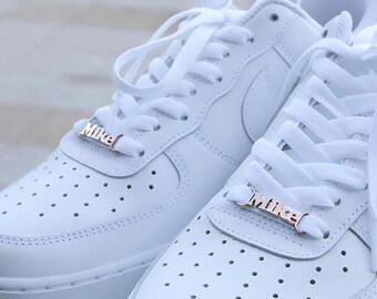 Puede incluir: Un par de zapatillas blancas de cuero con cordones blancos y etiquetas de nombre en oro rosa que dicen "Mike" en cada zapato.