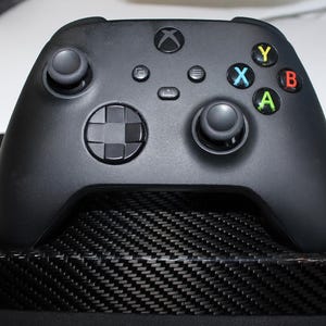 Puede incluir: Un mando negro de Xbox con botones de colores (Y, X, B, A) y una cruceta, apoyado en un soporte con textura de fibra de carbono. El mando tiene dos palancas analógicas y varios botones de función. El soporte es rectangular.