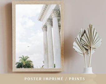 Op de afbeelding: Een ingelijste print met een klassieke architectuurscène met witte zuilen tegen een bewolkte lucht. De print zit in een licht houten frame. De tekst "POSTER IMPRIMÉ / PRINTS" staat onderaan de afbeelding.