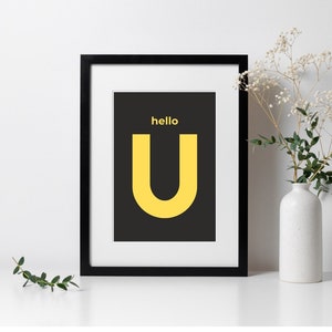 Puede incluir: Una impresión enmarcada en negro con una letra amarilla "U" sobre un fondo negro. El texto "hello" está por encima de la letra.