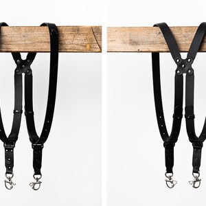 May include: Black leather camera straps with metal clips and adjustable buckles. The straps are hanging from a wooden beam.